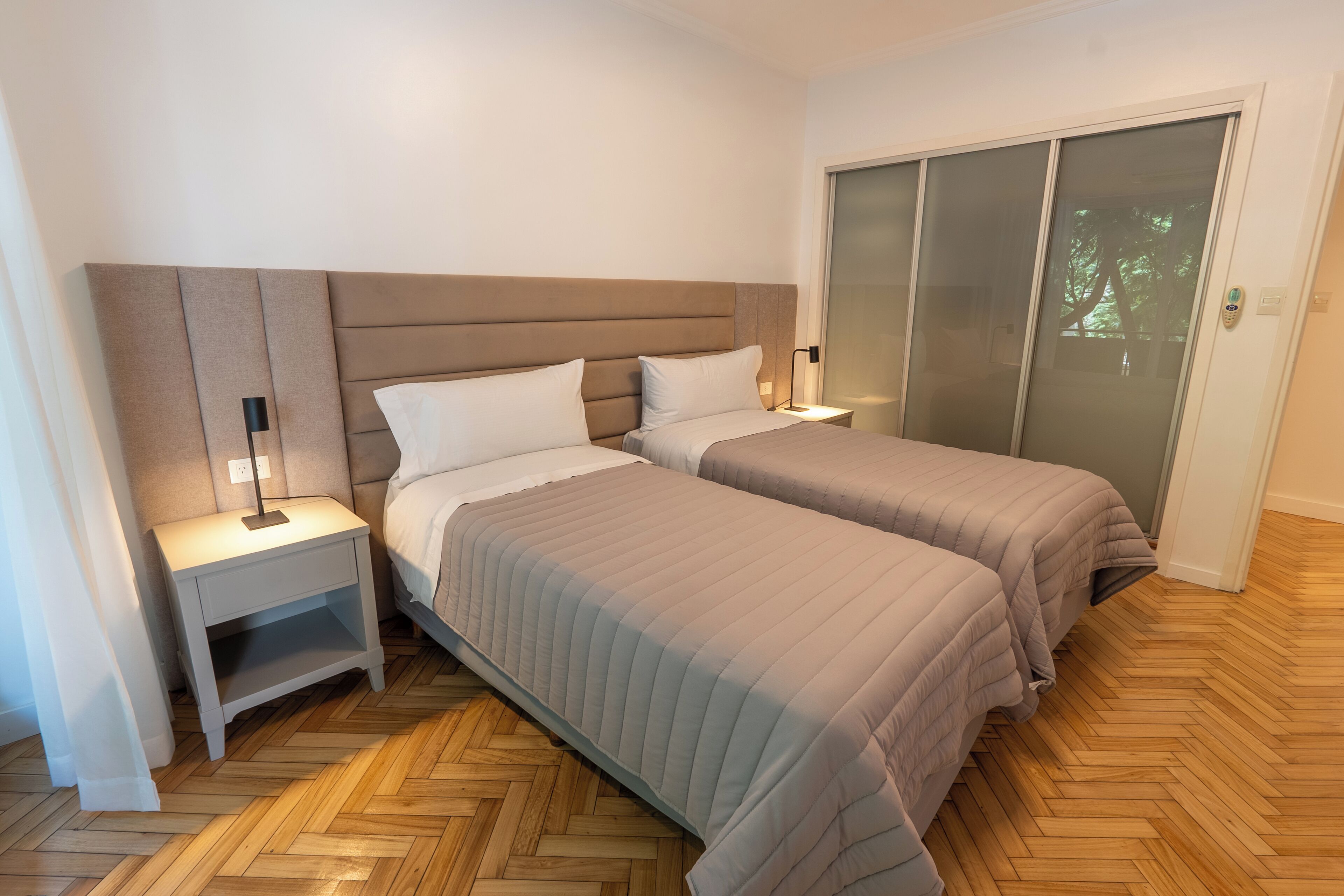 A bedroom with two beds, a nightstand with a lamp, and a sliding glass door.