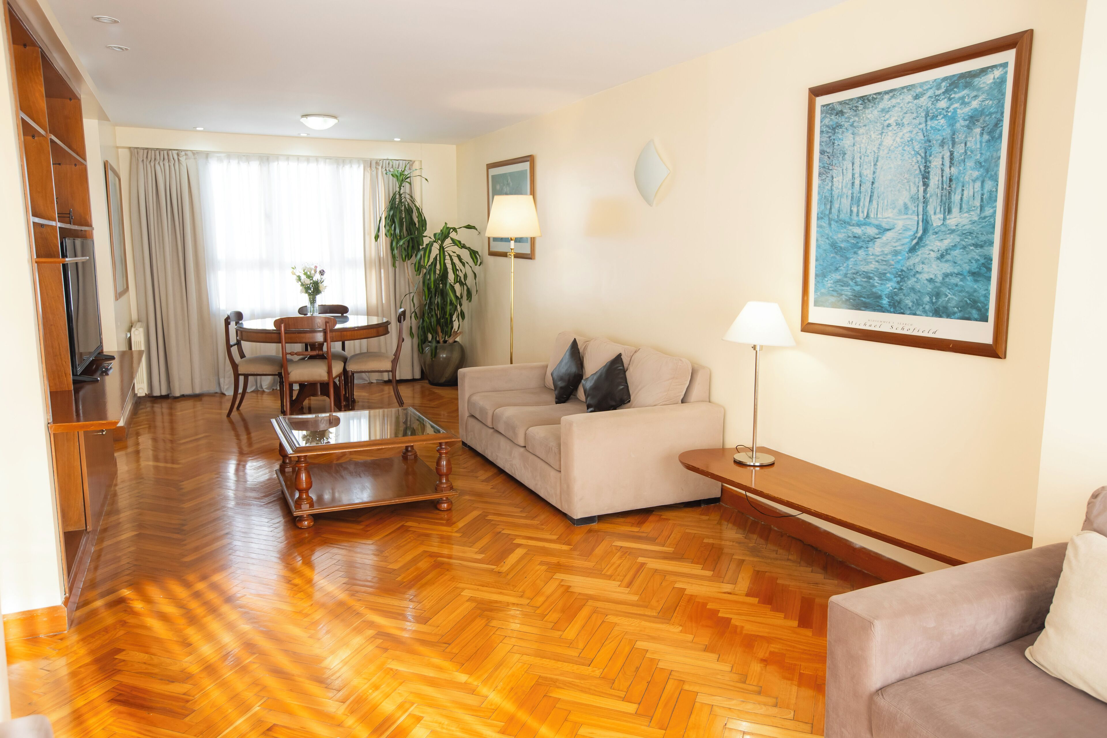Una sala de estar con sofá, una mesa de centro, lámpara, un cuadro y un comedor con mesa y sillas.
