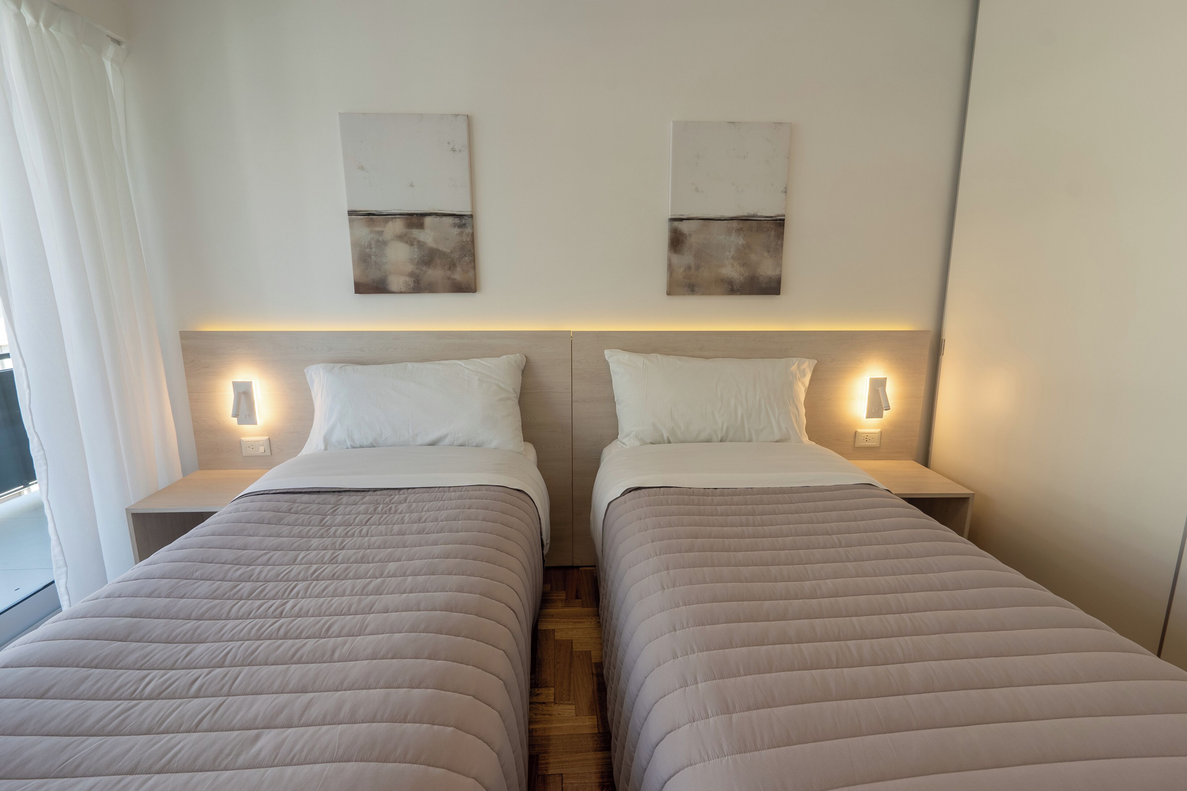 A hotel room with two beds, each with a headboard, bedside tables, and wall-mounted light fixtures.