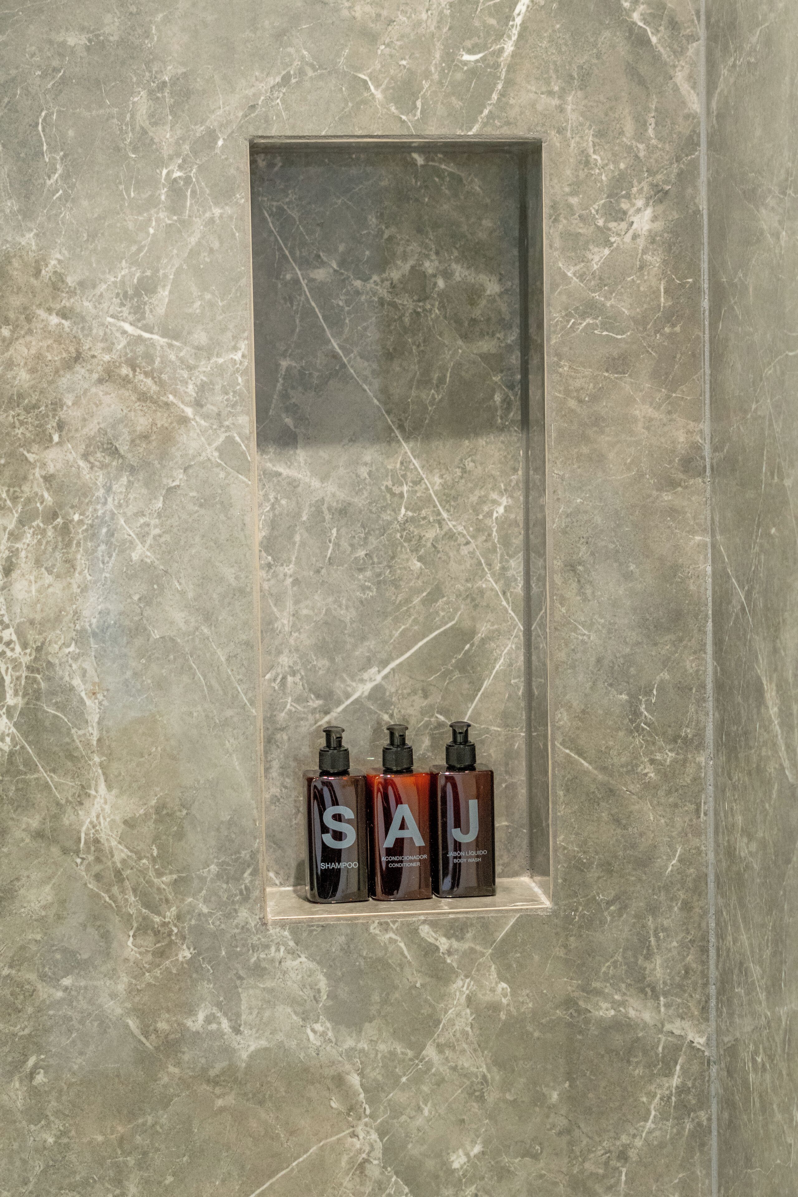 Un baño con pared de mármol y repisa que contiene tres botellas etiquetadas como "SAJ SHAMPOO" y "SAJ LIQUIDO".