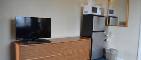 A room with a wooden dresser, a television, a microwave, a refrigerator, a trash can, and a wall-mounted mirror.