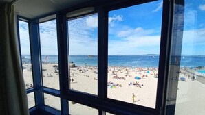 A view from inside a room with large windows overlooking a crowded beach with people and boats.