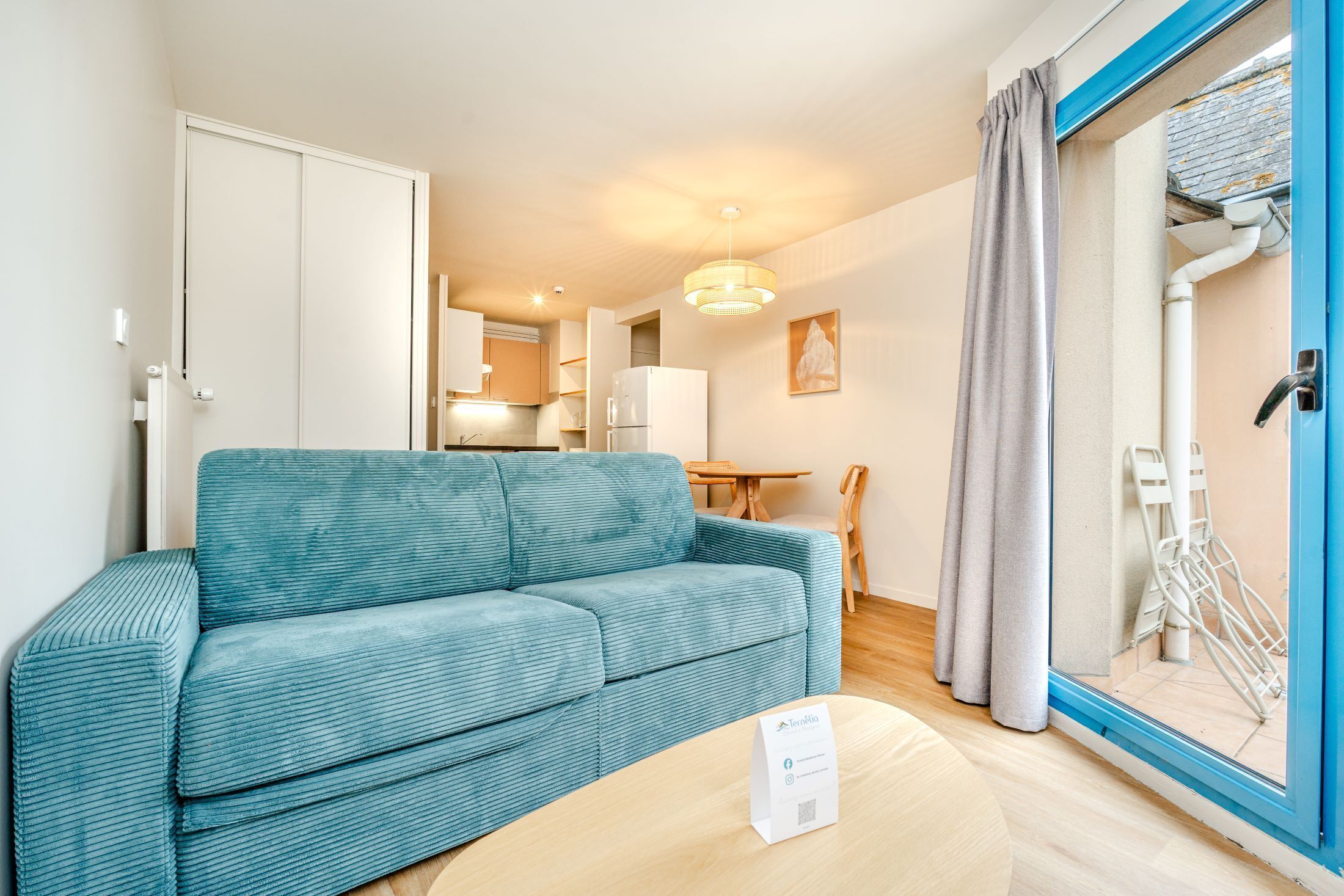 A room with a sofa, a table, a door, a window with curtains, and a view of a kitchen area.