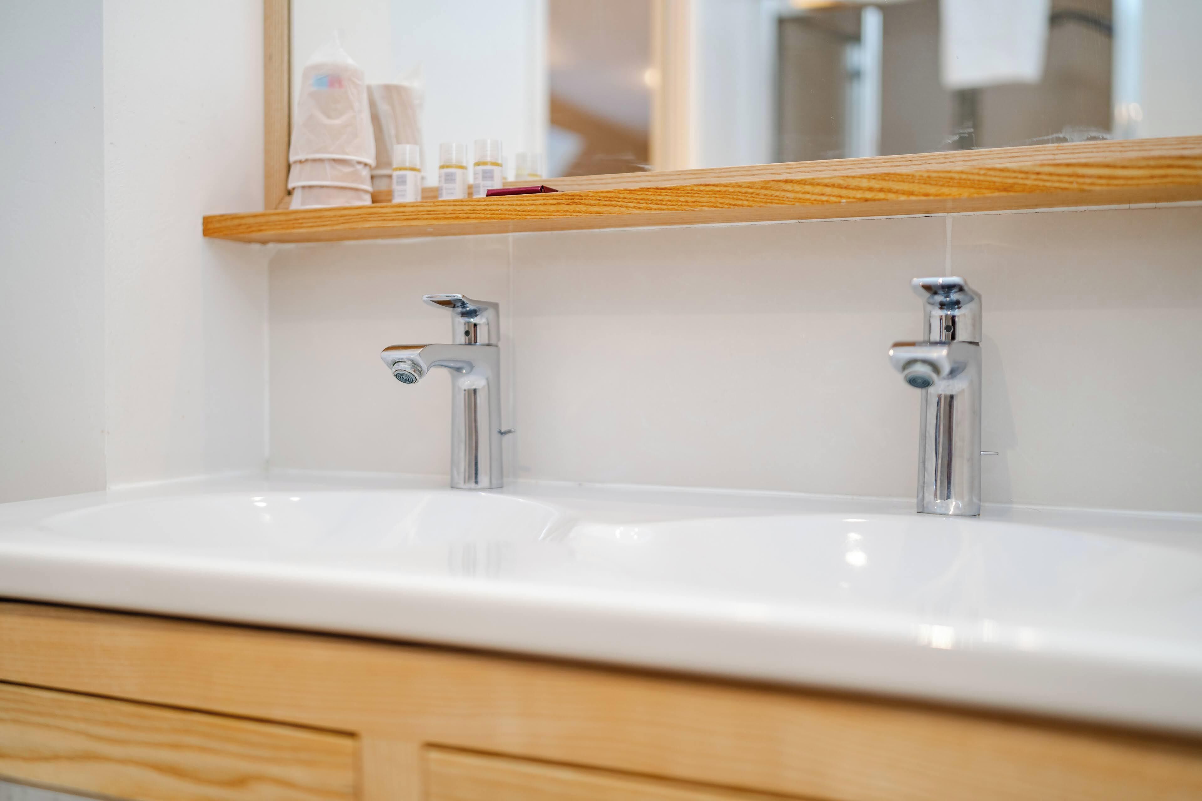 A bathroom with a white sink, two faucets, and a wooden shelf above the sink.
