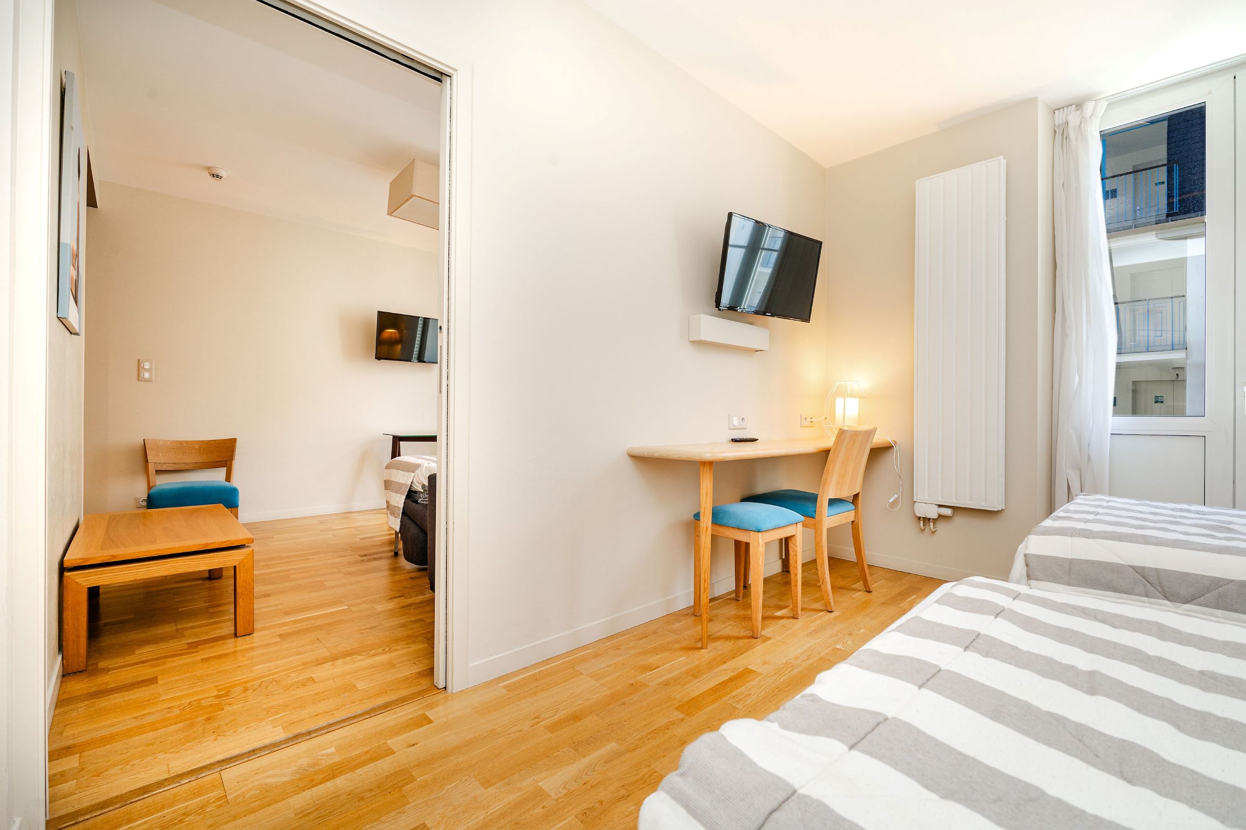 A hotel room with a wooden floor, a bed with striped bedding, a desk with a chair, a television mounted on the wall, and a window with curtains.