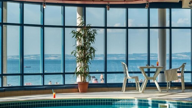 An indoor swimming pool with a large window view of the sea, a potted plant, and a table with chairs.