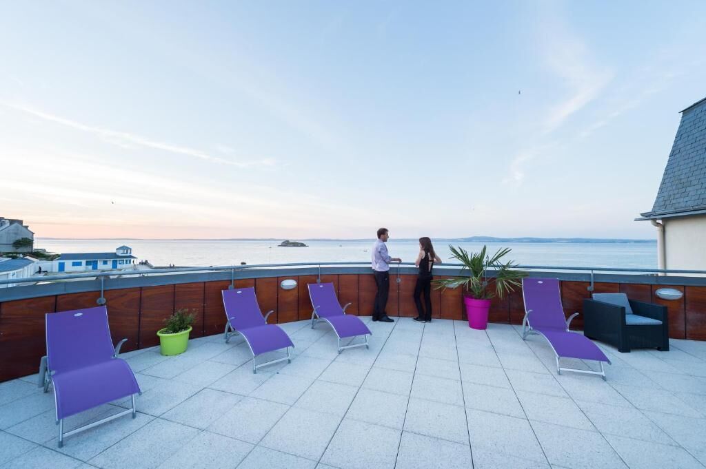 A rooftop terrace with lounge chairs, potted plants, and a view of the sea.