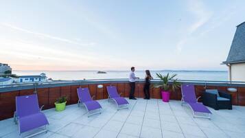 A rooftop terrace with lounge chairs, potted plants, and a view of the sea.