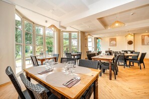 A dining area with multiple tables and chairs, large windows, and hanging light fixtures. - Les Résidences d'Armor (Douarnenez)