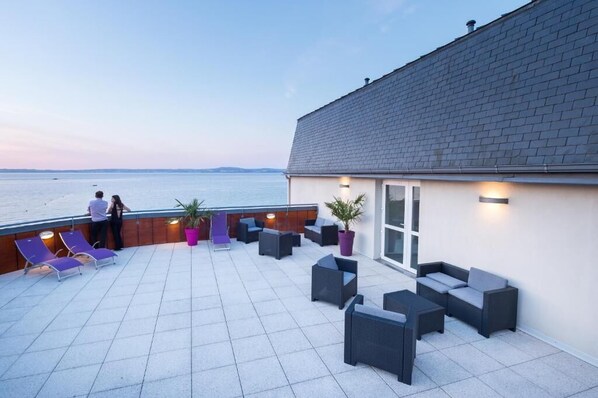 A rooftop terrace with outdoor seating, a view of the sea, and a building with a tiled roof. - Les Résidences d'Armor (Douarnenez)