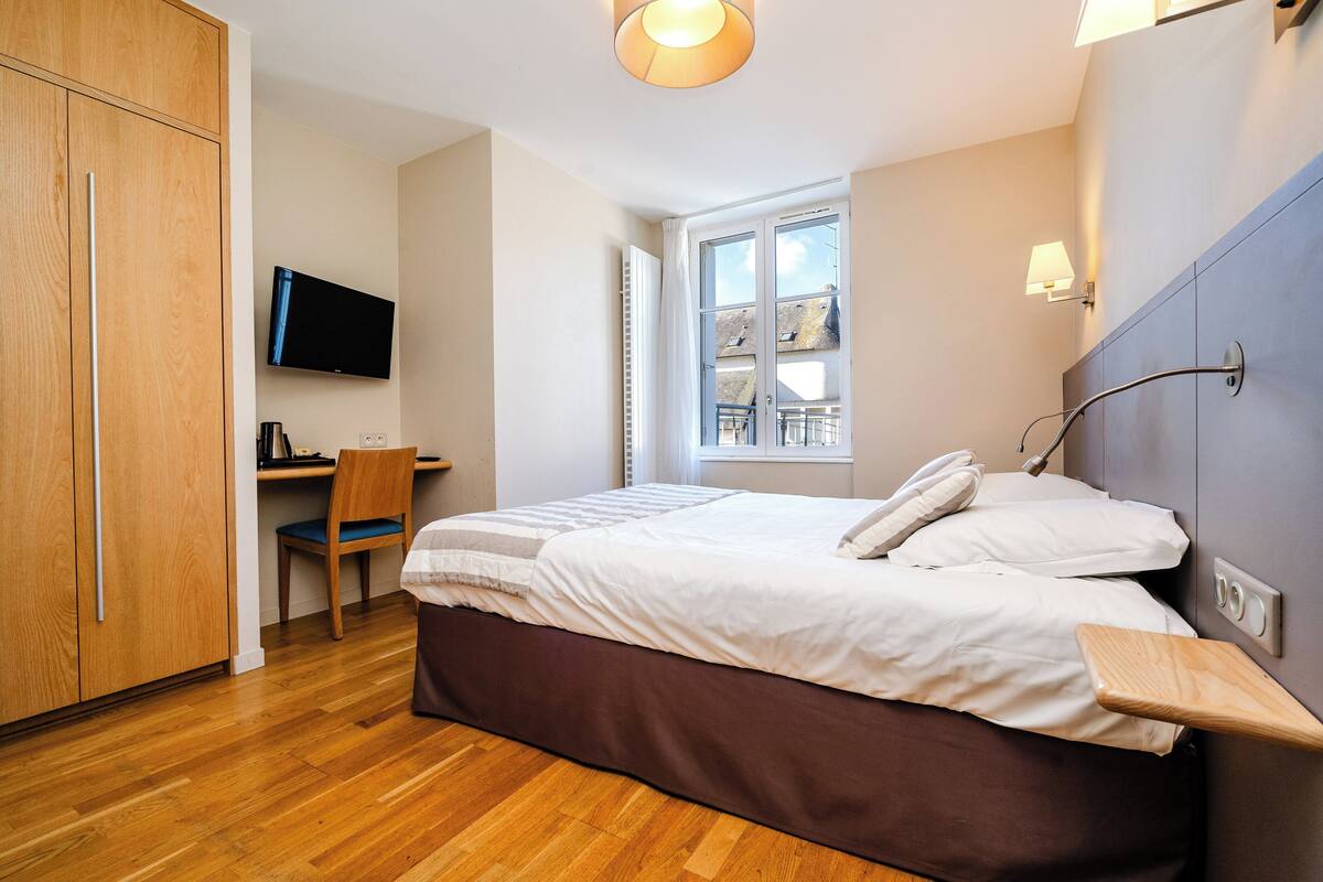 A bedroom with a bed, a desk, a chair, a TV, and a window.