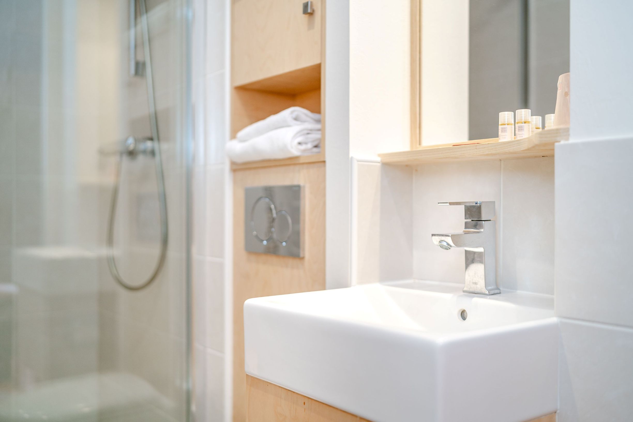 A modern bathroom with a glass shower enclosure, a white sink, and wooden shelving with toiletries.
