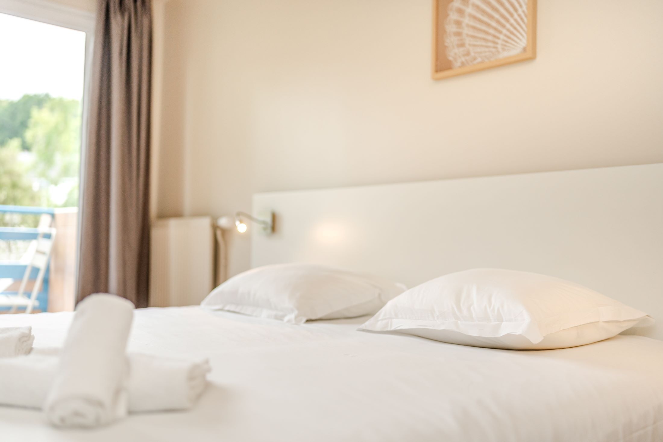 Un lit bien fait, avec du linge de lit blanc et des oreillers, une lampe de chevet et une vue sur la verdure à l’extérieur.