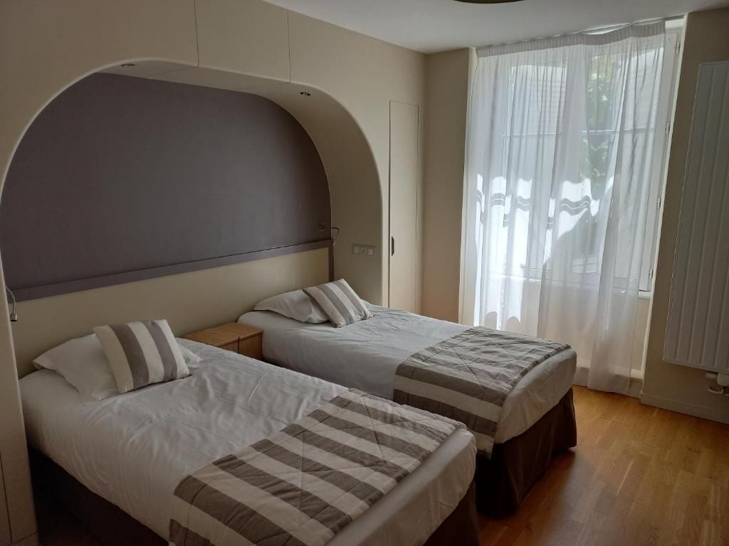 Une chambre avec deux lits, une tête de lit incurvée, une fenêtre avec des rideaux légers et un plancher en bois.
