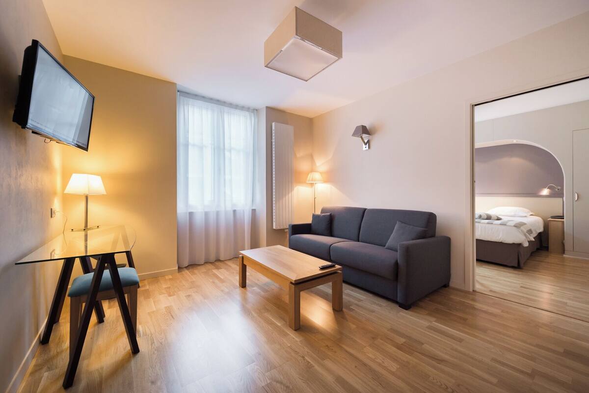 A modern hotel room with a sofa, a coffee table, a TV mounted on the wall, and a bed in the adjacent room.