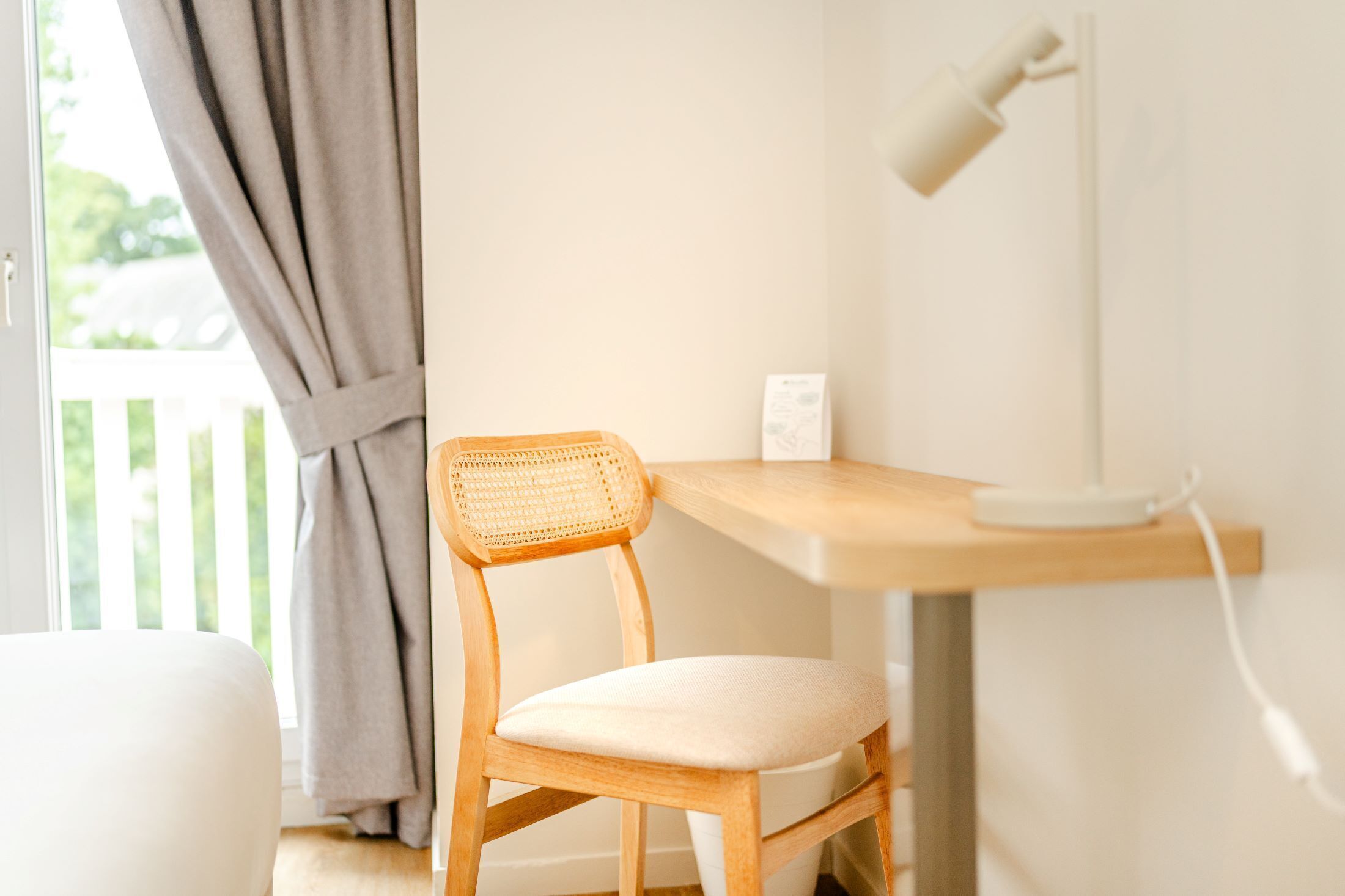 Una silla de madera con respaldo trenzado y asiento acolchado, un escritorio blanco con lámpara, y una pared blanca.