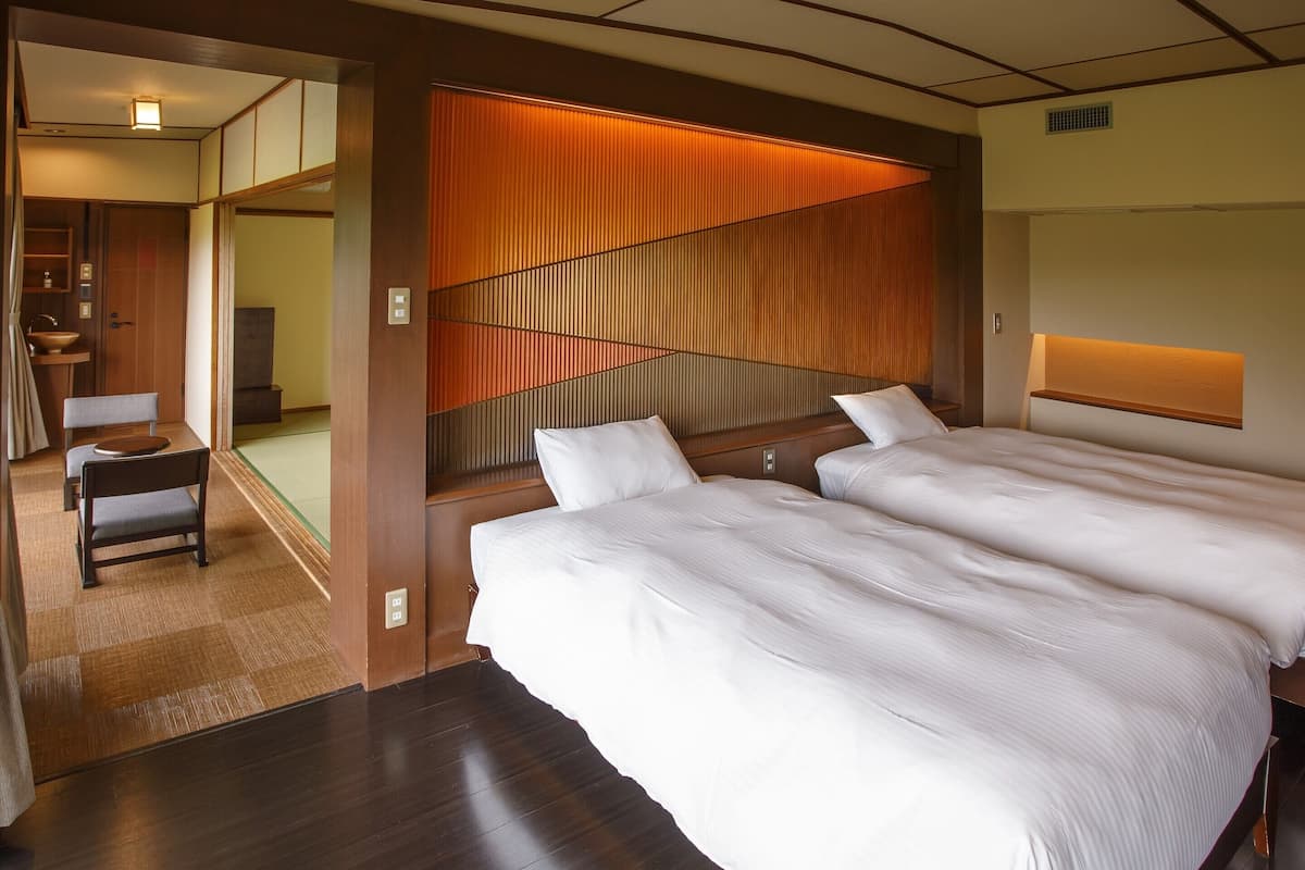 Habitación de hotel con dos camas, cabecero de madera, una mesita con una silla y vistas a una habitación tradicional japonesa.