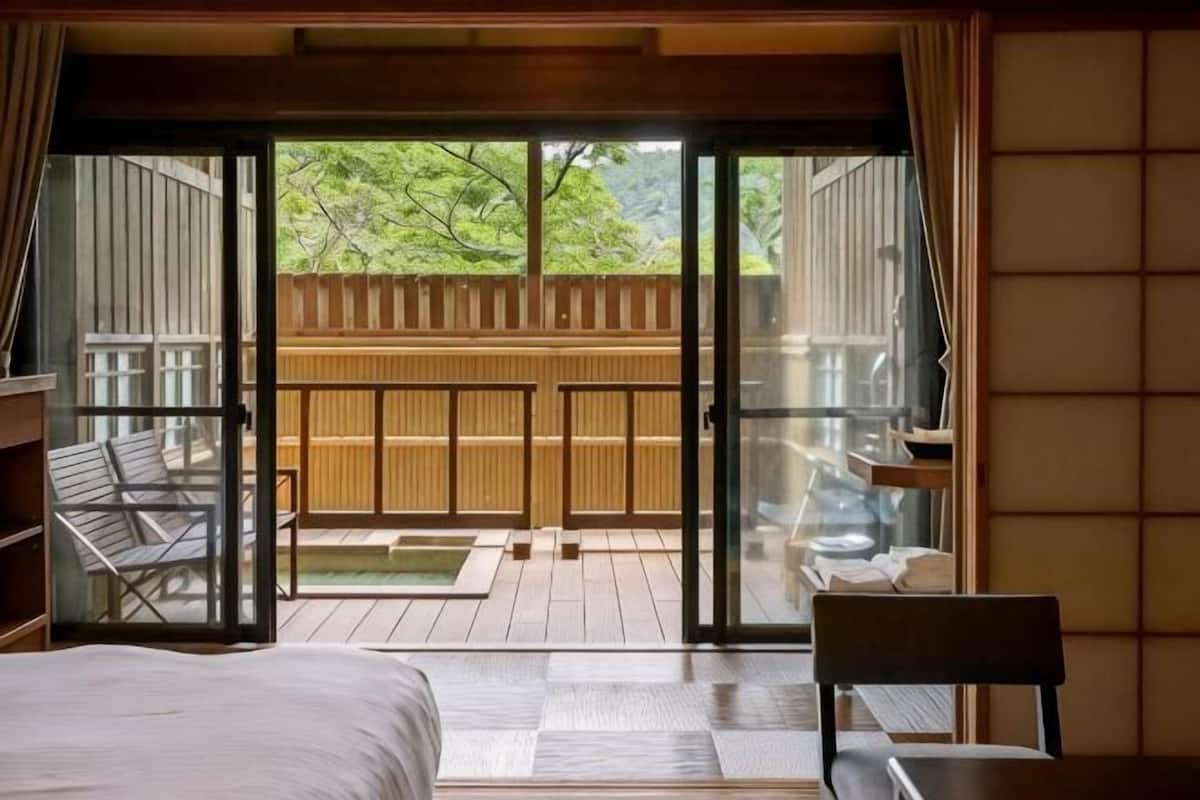 Una habitación con cama, una silla, una mesa y un ventanal corredera que da a un balcón con cerramiento de madera y vistas a árboles.