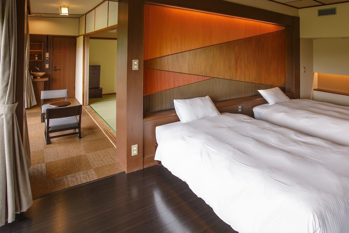 A hotel room with two beds, a wooden headboard, a small seating area with a chair and table, and a view of a traditional Japanese room.
