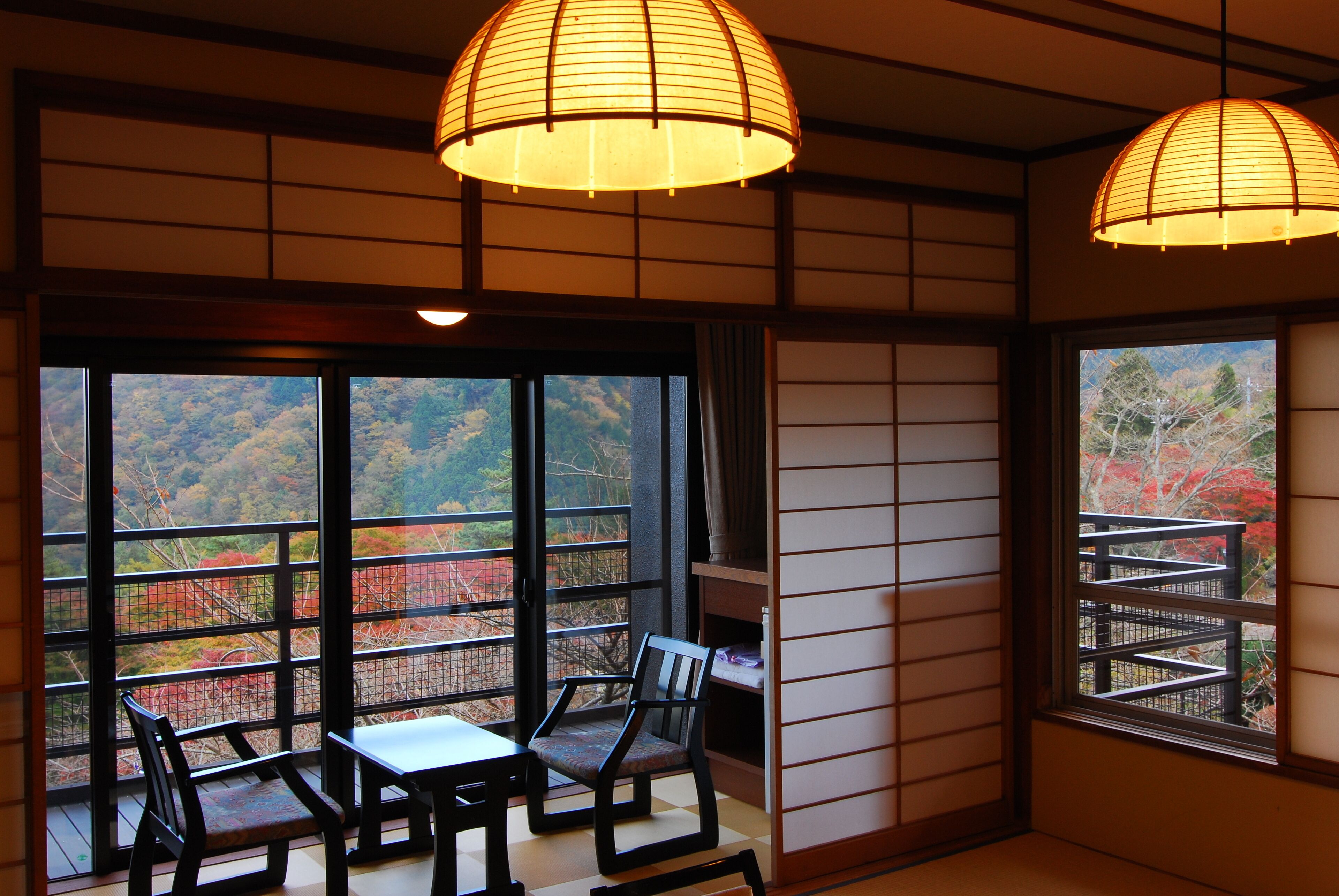 Ein traditioneller japanischer Raum mit Tatami-Bodenbelag, einem niedrigen Tisch, zwei Stühlen und großen Fenstern mit Blick auf Herbstlaub.
