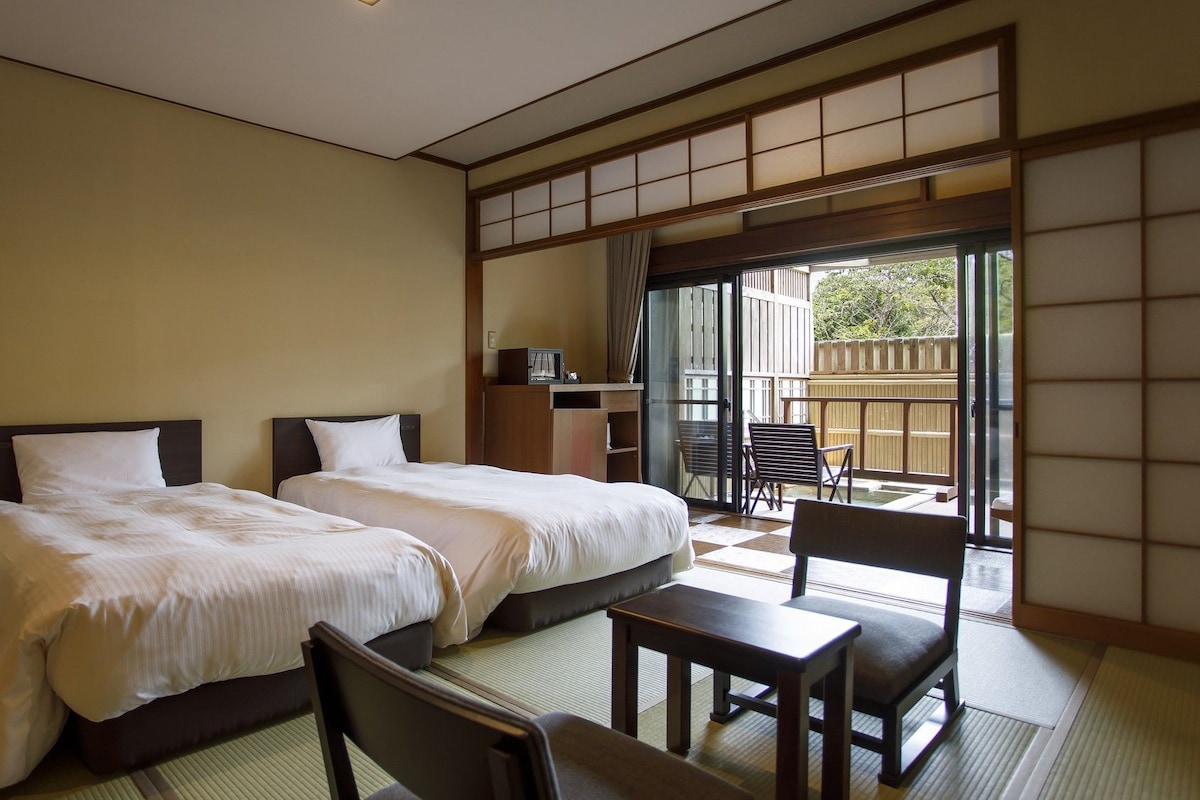A hotel room with two beds, a small table, and a chair. There is a sliding door leading to an outdoor area with a view of trees.