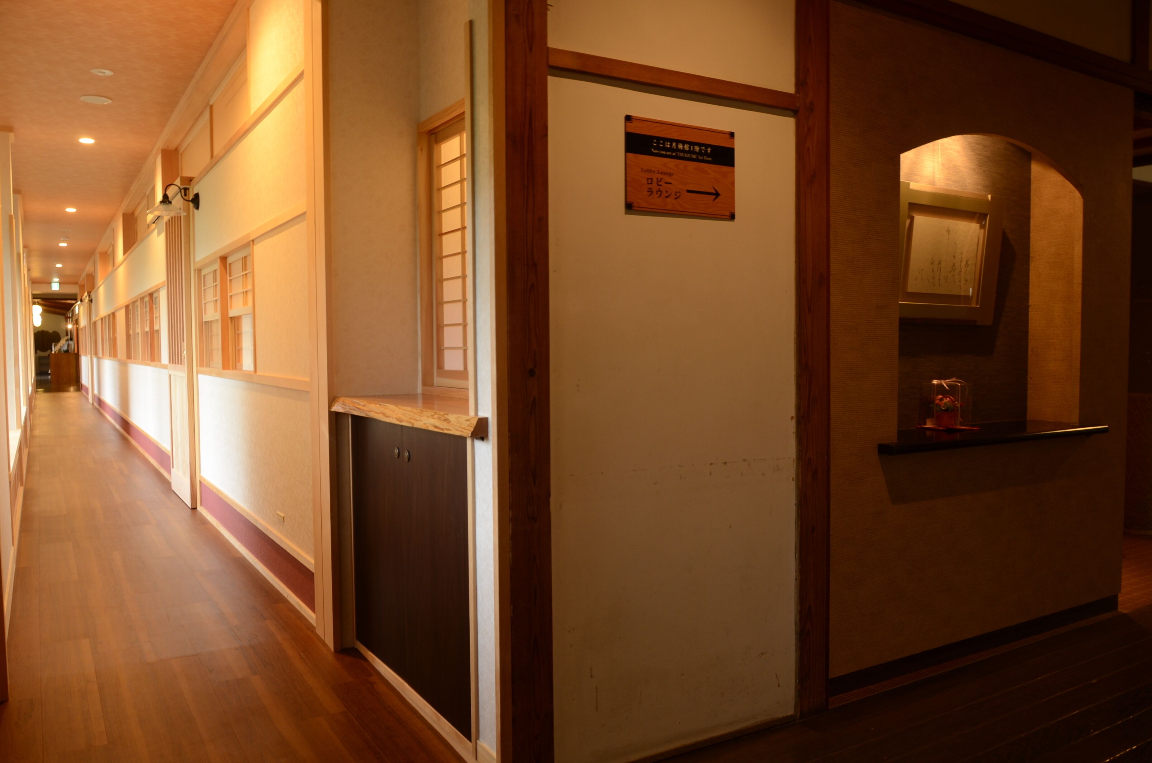 A hotel corridor with wooden flooring, a reception desk, and a sign indicating the direction to the lobby.