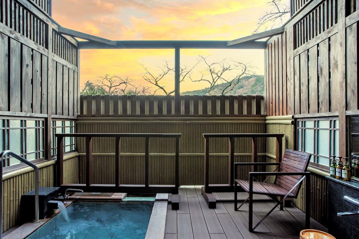 Una terraza de madera con jacuzzi, asientos y vistas a un atardecer.