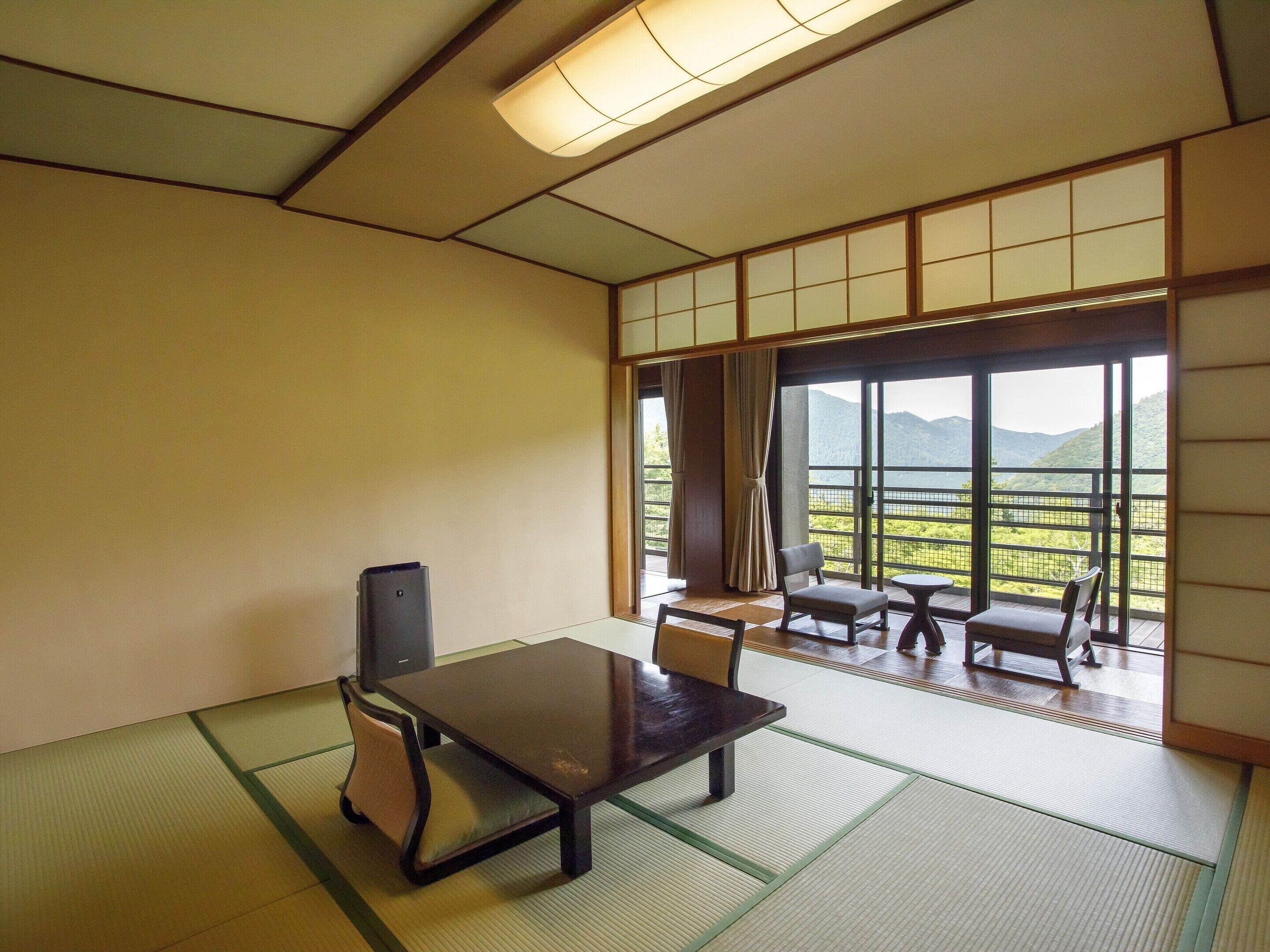 A traditional Japanese-style room with tatami flooring, a low table, and chairs. A large window offers a view of a mountainous landscape.