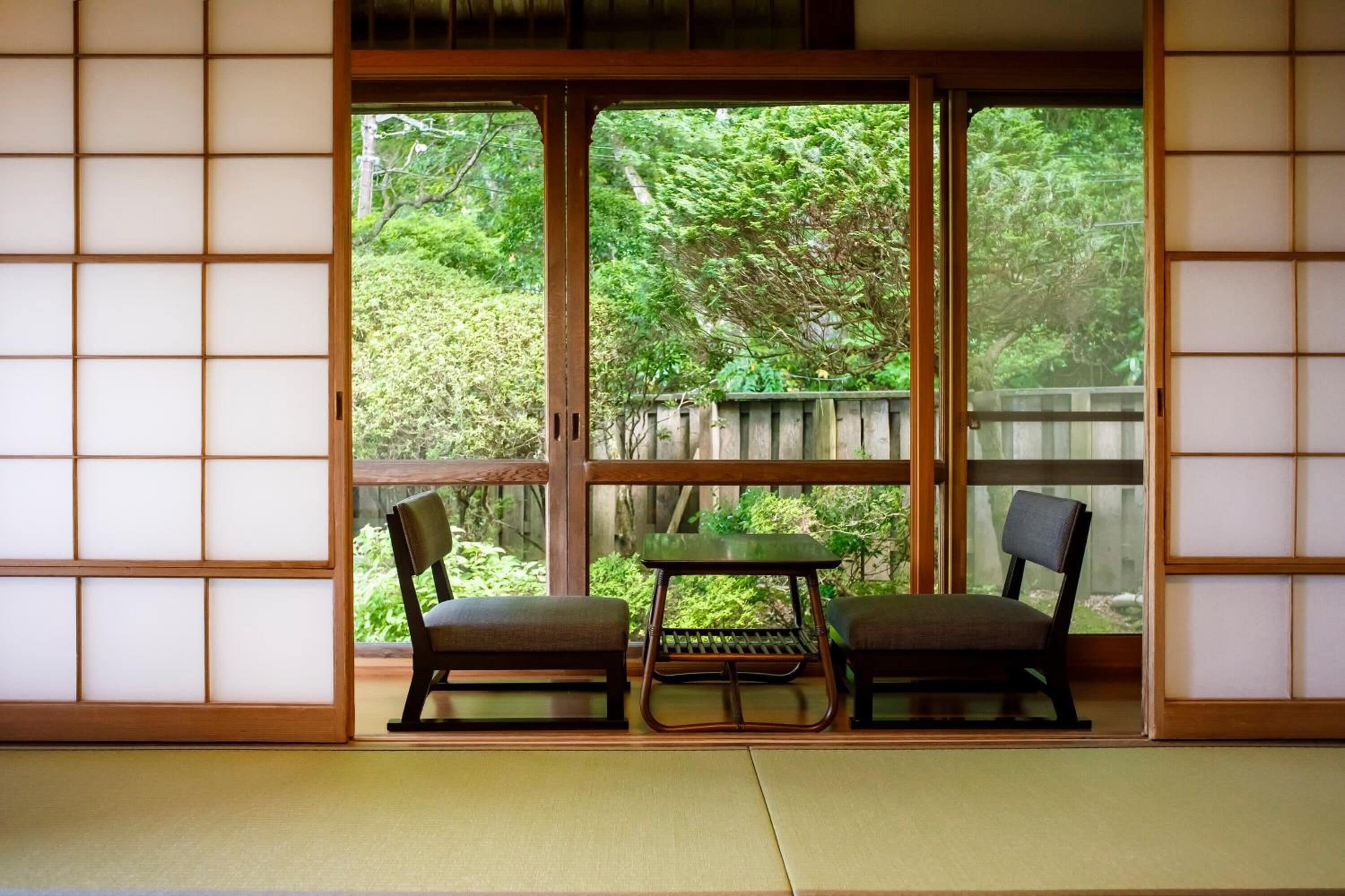 A traditional Japanese room with tatami flooring, a sliding shoji screen, and a view of a wooden fence and greenery outside.