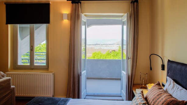 A bedroom with a bed, a window with curtains, a radiator, and a view of greenery outside. - Aethra Rooms (Nafplio)