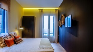 A modern bedroom with a large bed, a bedside lamp, and a view of the outside through a window with curtains. - Aethra Rooms (Nafplio)