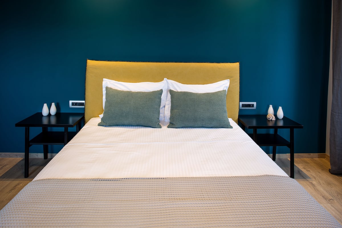 A neatly made bed with a white duvet and two green pillows, flanked by black nightstands with white vases.