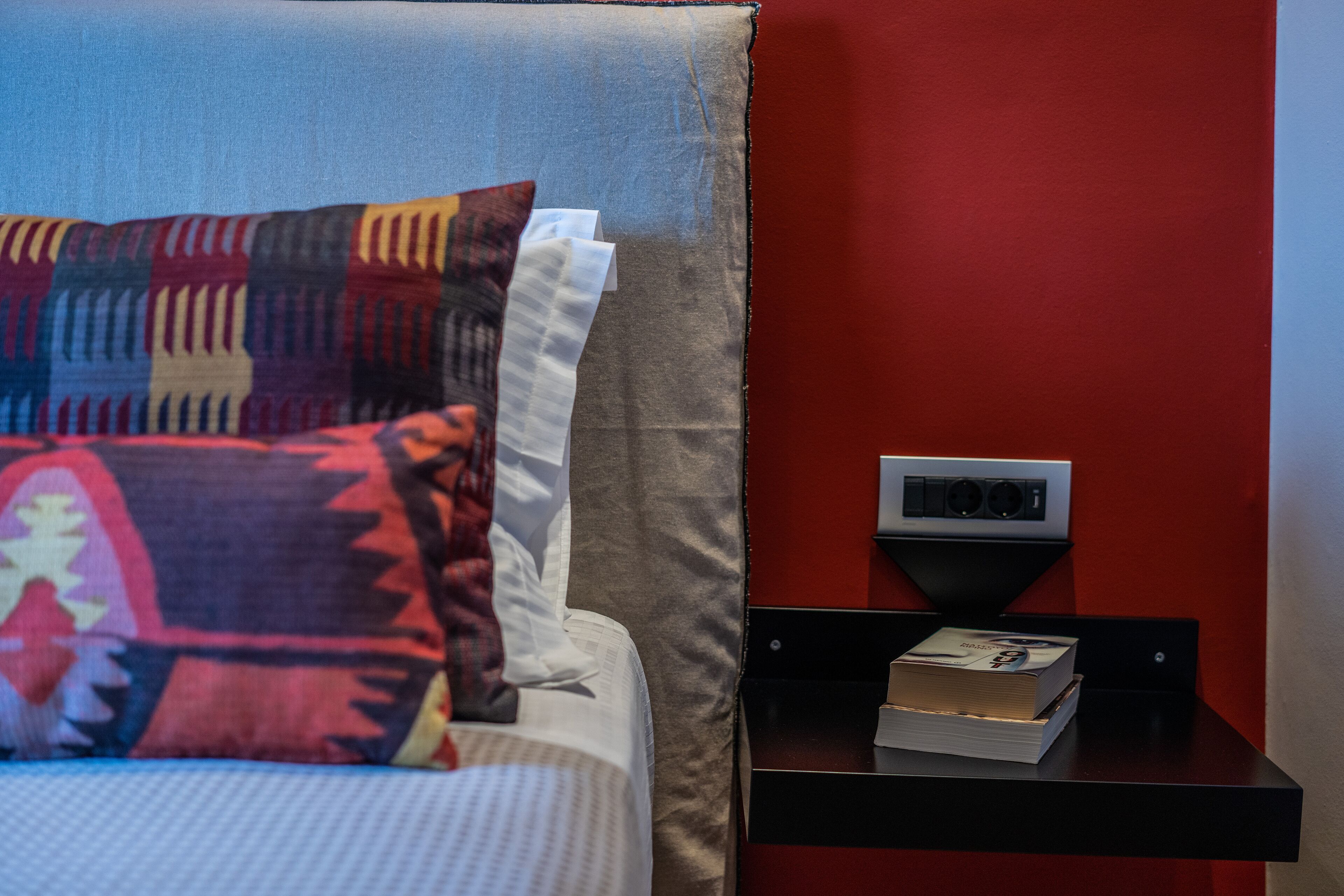 A bed with patterned pillows, a bedside table with a book, and a wall-mounted device.