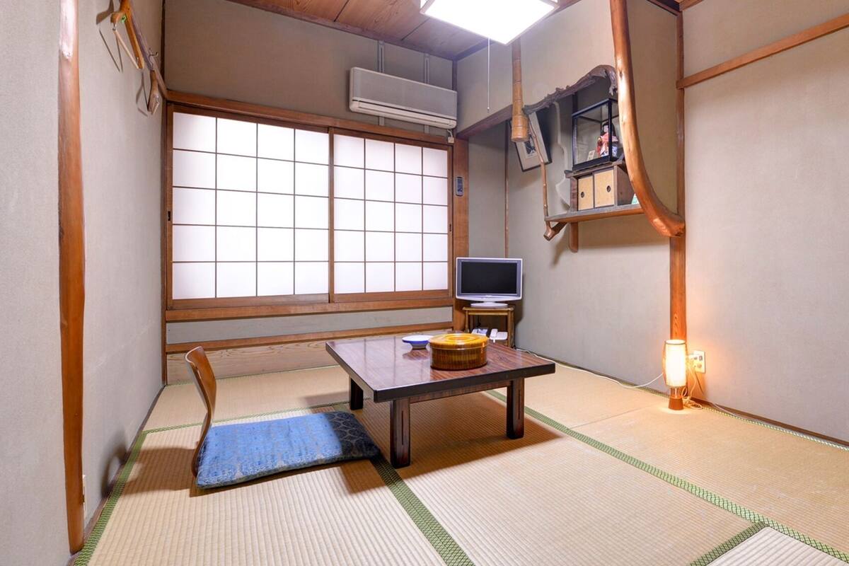 A traditional Japanese room with tatami flooring, a tatami mat, a low table with a television, and a shelf with items.