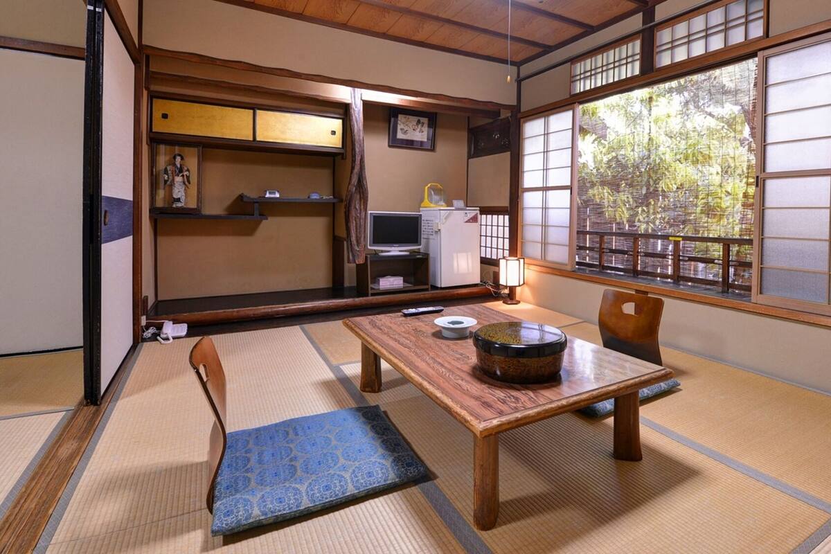 A traditional Japanese room with tatami flooring, a low table with a bowl, a chair, a TV, and a refrigerator.