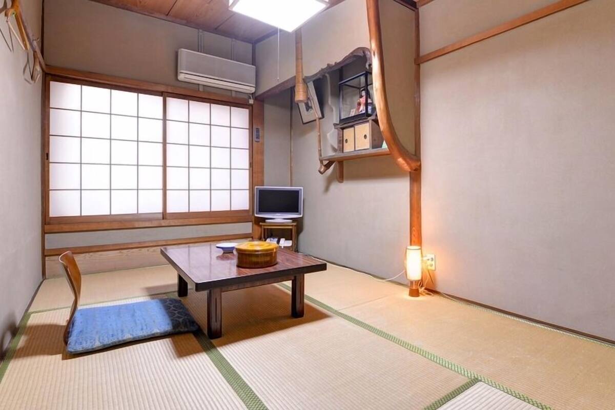 A traditional Japanese room with tatami flooring, a tatami mat, a television on a low table, and a shelf with items.