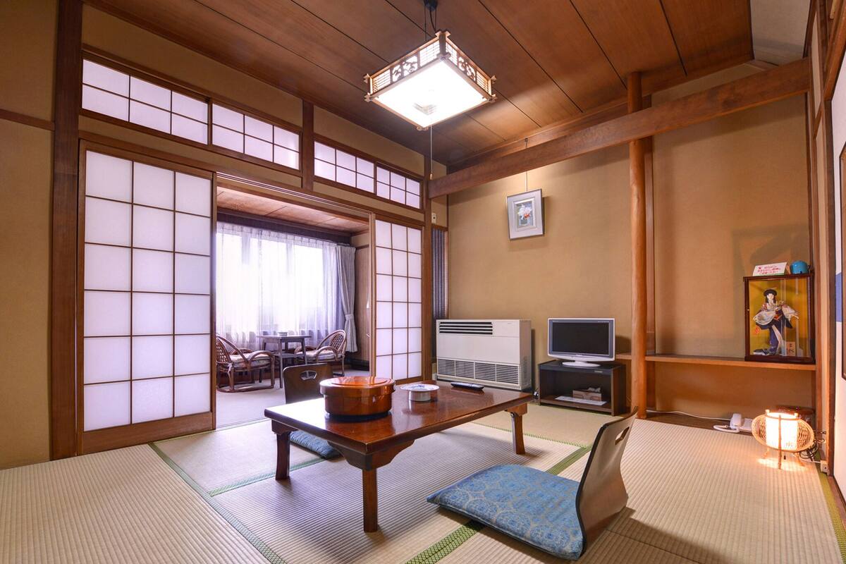 A traditional Japanese room with tatami flooring, a low table with a cushion, a television, and a heater.