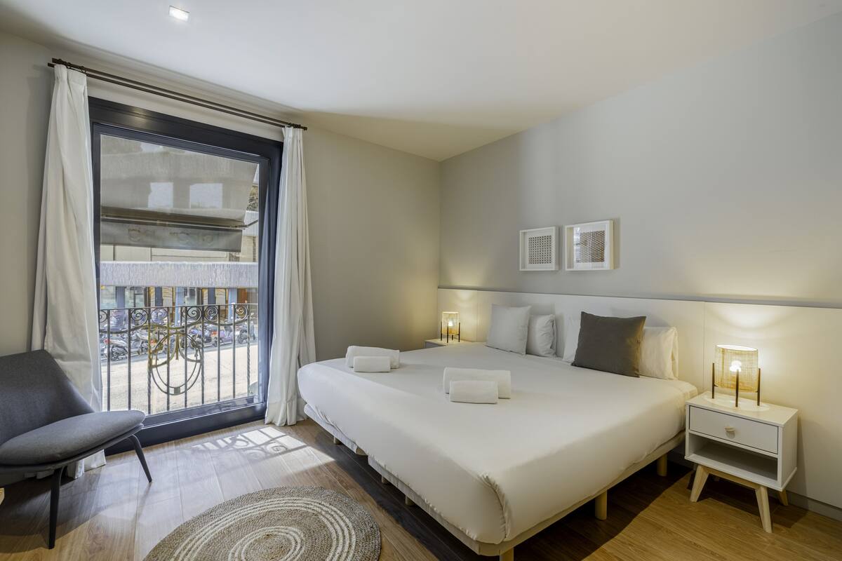 A modern hotel room with a large bed, bedside tables, a chair, and a view of the outside.