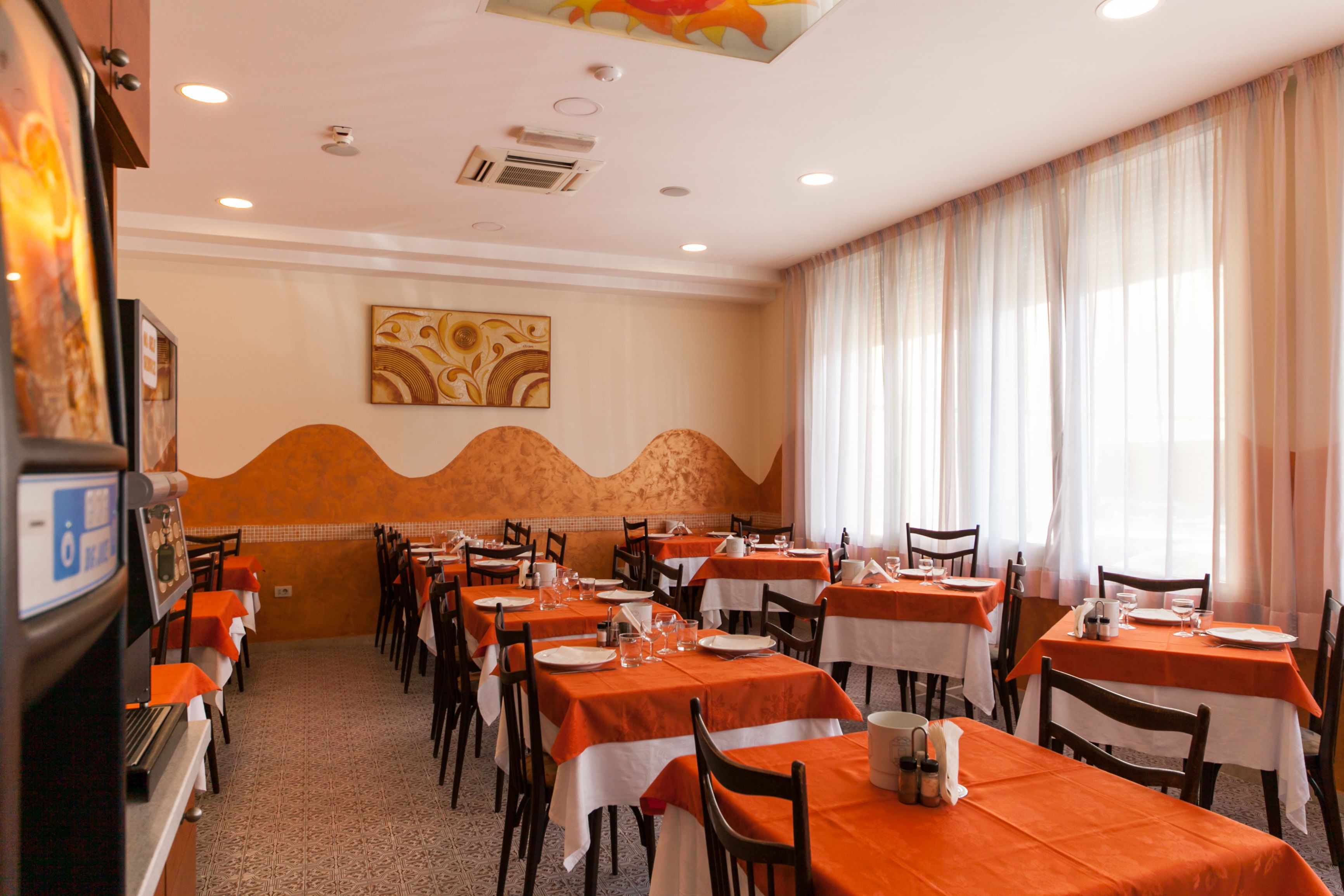 A dining area with tables set for meals, a large window with curtains, and a wall with a decorative pattern.