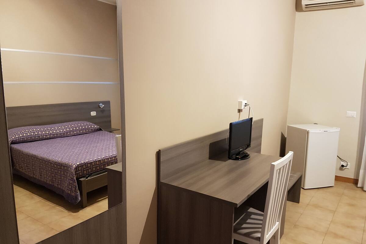 A bedroom with a bed, a desk, a chair, a small white cabinet, and an air conditioner.