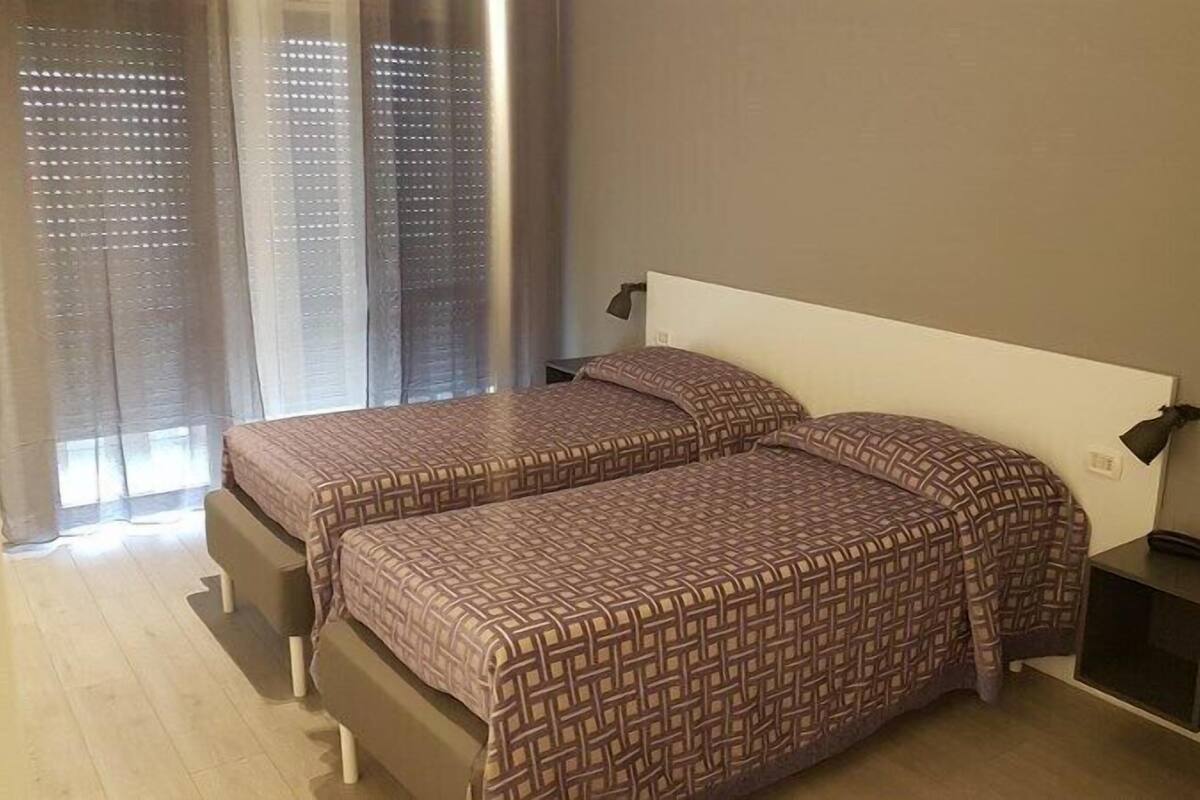 A bedroom with two beds, a nightstand, a lamp, and a window with curtains.