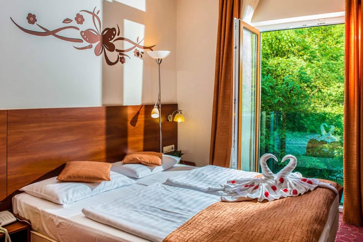 A hotel room with a large bed, two bedside lamps, a floral wall decal, and a view of greenery through the window.