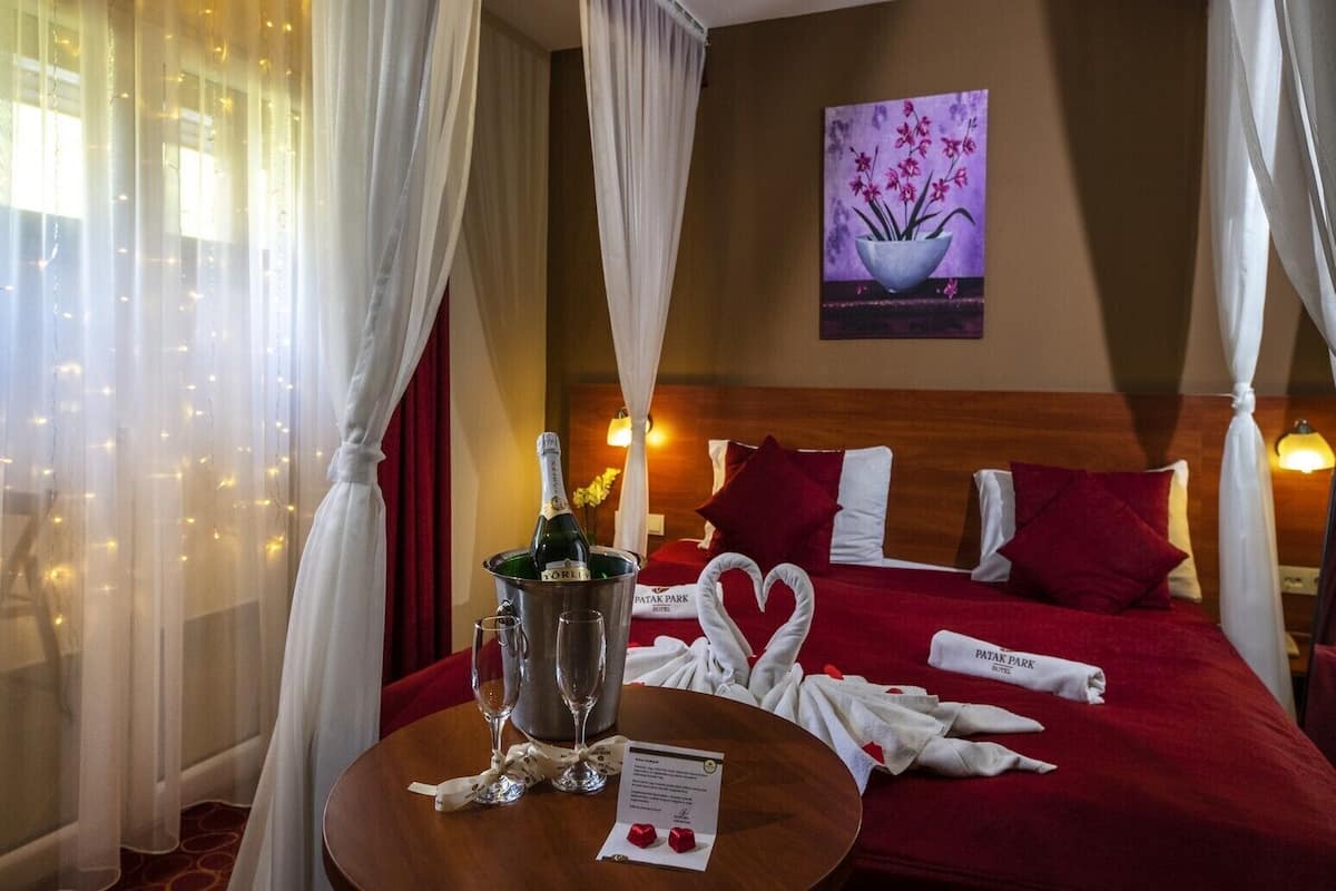 A hotel room with a bed, a bedside table with a bottle of champagne, two glasses, and a heart-shaped towel arrangement.