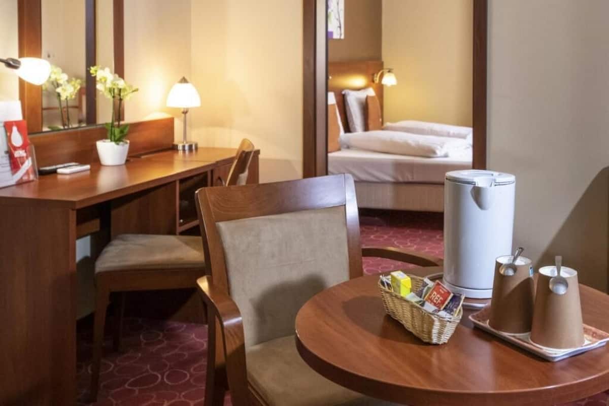 A hotel room with a wooden desk, a chair, a round table with a basket and two paper cups, a bed with white linens, a bedside lamp, and a large white appliance.