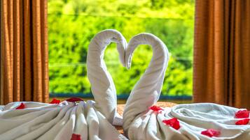 Una habitación con vistas a la vegetación por una ventana, una toalla dispuesta en forma de corazón y una cama con sábanas blancas y pétalos de rosa roja.