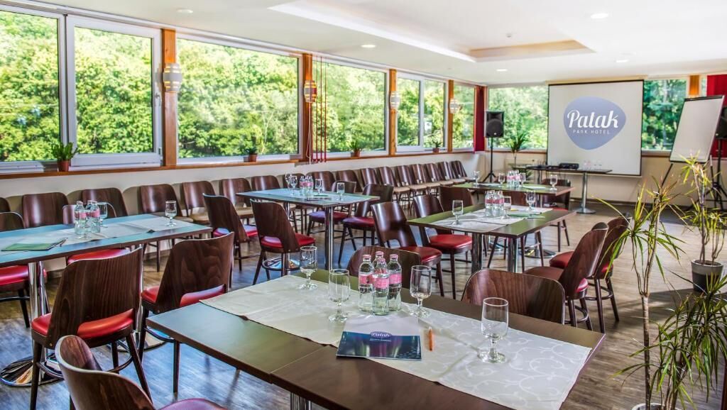 Een vergaderzaal met meerdere tafels en stoelen, grote ramen met uitzicht op groen, en een scherm waarop de naam van het hotel staat.