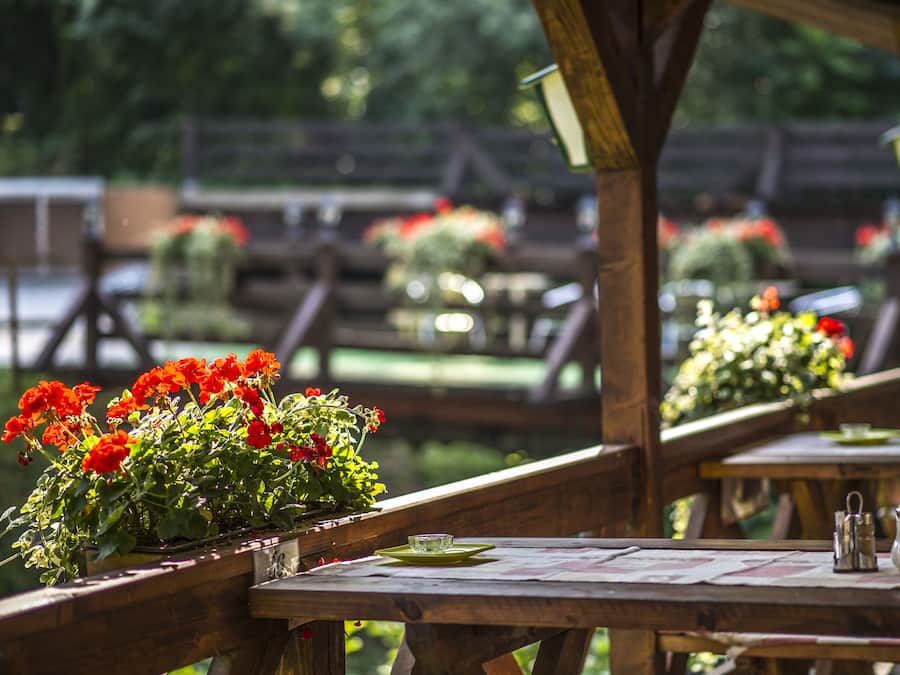 A wooden outdoor dining area with tables and benches, surrounded by lush greenery and red flowers.