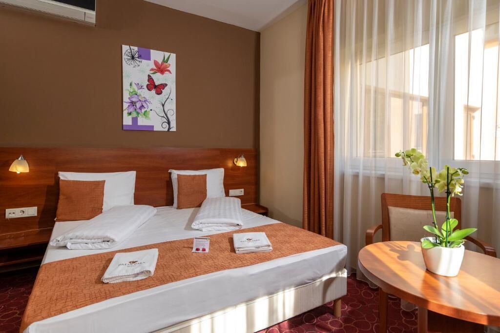 A hotel room with a large bed, two bedside tables, a small table with a plant, and a painting on the wall.