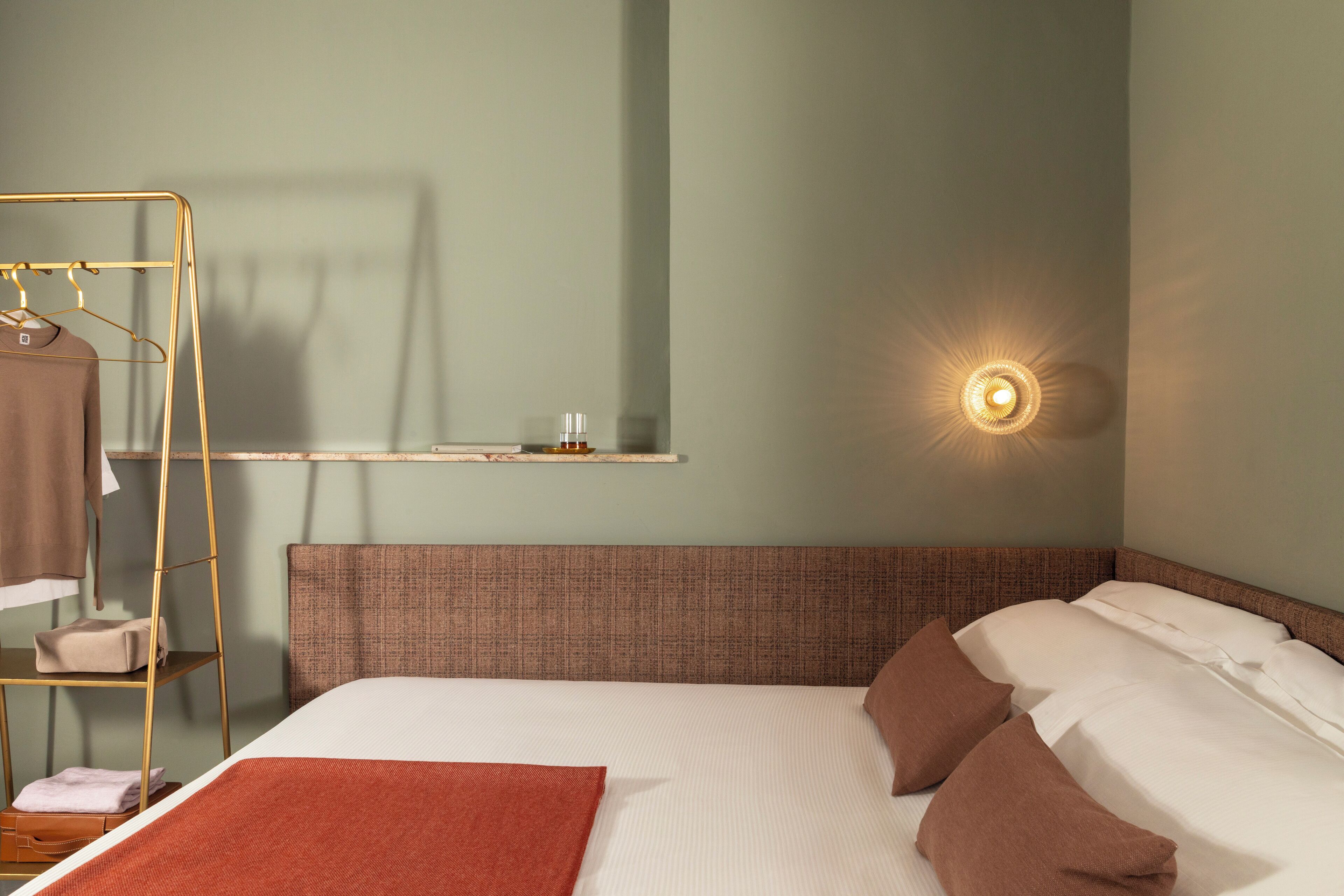 A hotel room with a bed, a hanging wardrobe, a shelf with a bottle, and a wall-mounted light.