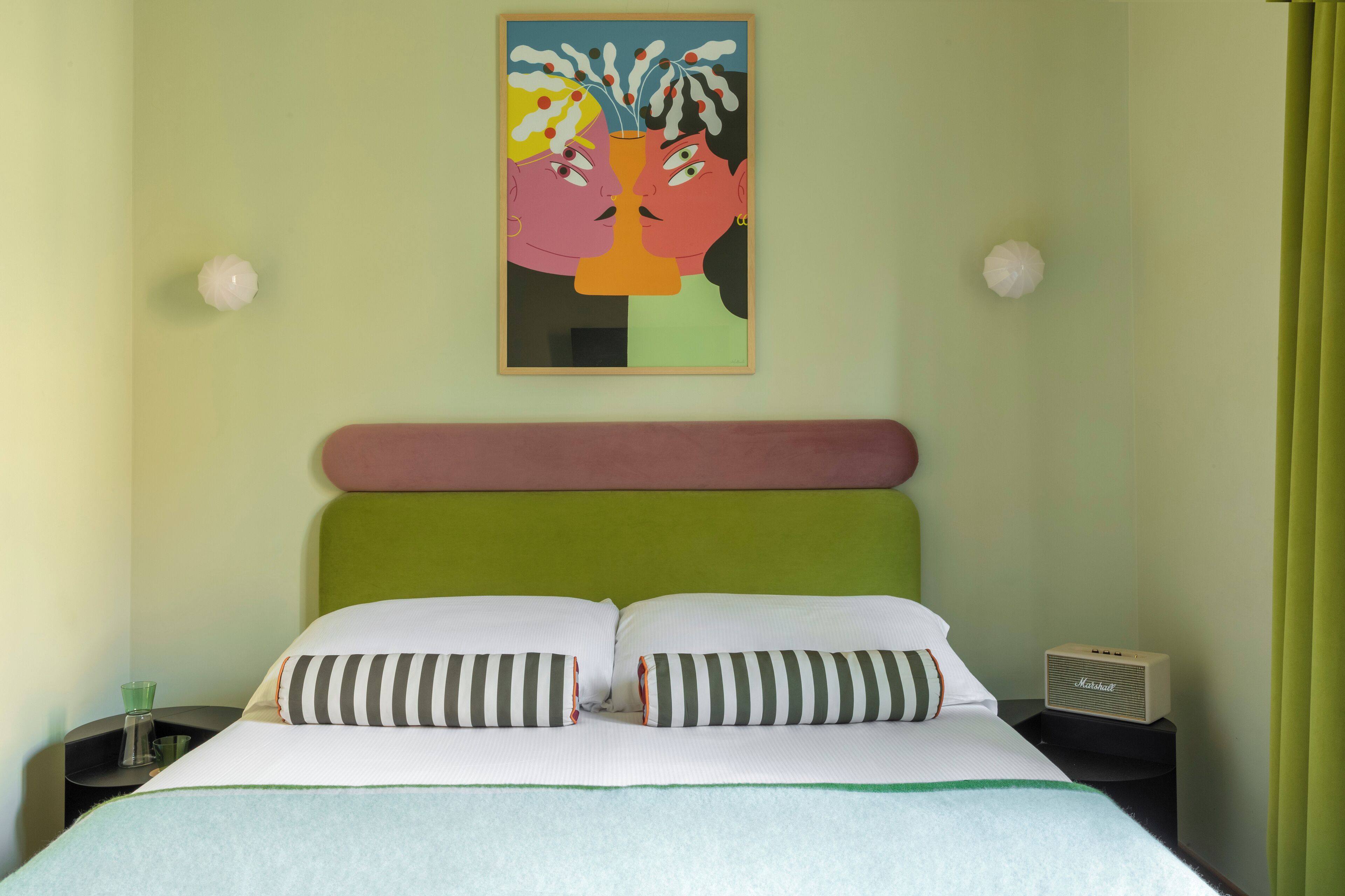A hotel room with a bed, striped pillows, a green headboard, a framed artwork on the wall, and a Marshall radio on a bedside table.